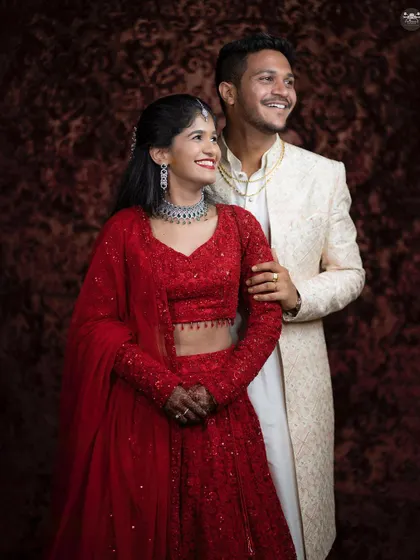 A happy, candid portrait of a couple sharing a laugh. The bride is wearing a vibrant red lehenga and diamond jewelry, while the groom is in a classic ivory sherwani.