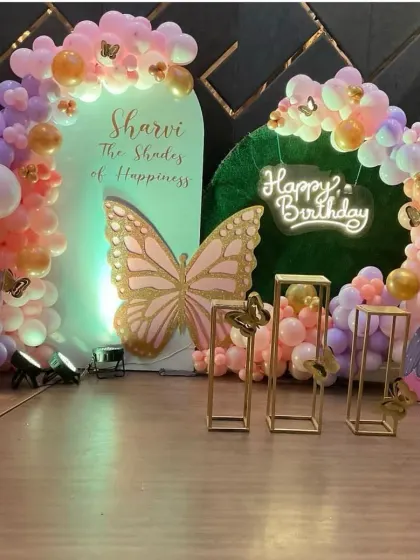A spectacular butterfly themed first birthday celebration. This setup includes a large, glittery butterfly centerpiece, a light-up marquee number '1', and a beautiful balloon arch in pastel shades of purple, pink, and orange.