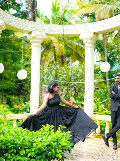 A beautiful pre-wedding portrait in a lush green setting. The couple is framed by a classic white gazebo, adding a touch of elegance.