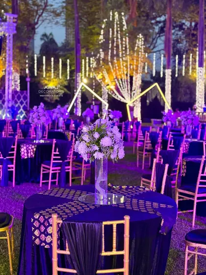 Elegant guest seating for a Sangeet, with deep blue tablecloths, gold-patterned runners, and simple floral centerpieces under a canopy of lights.