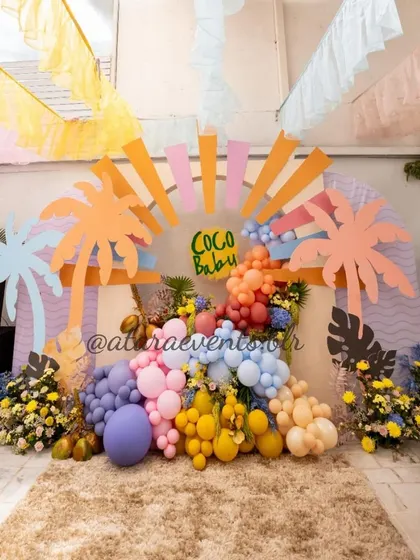 A vibrant and tropical "Coco Baby" themed baby shower. The backdrop is a burst of color with palm tree cutouts, a sun motif, and a lush arrangement of balloons and flowers.