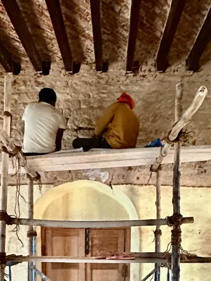 Two workers sit on a simple wooden scaffold, repairing the upper portion of a wall. This image speaks to the quiet, focused labor that goes into restoring a heritage building.