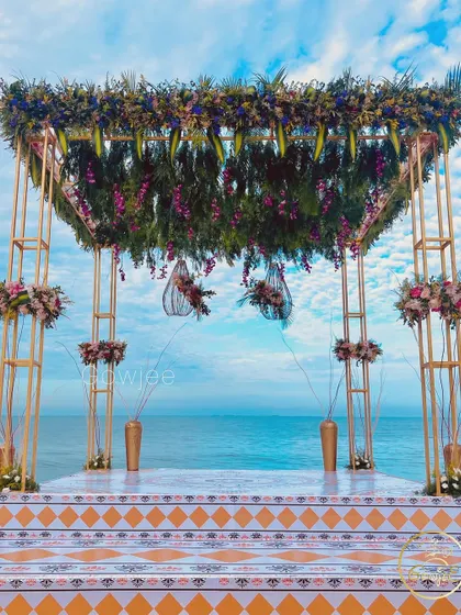 A breathtaking beach wedding mandap with a lush canopy of greenery and purple flowers against the blue ocean.