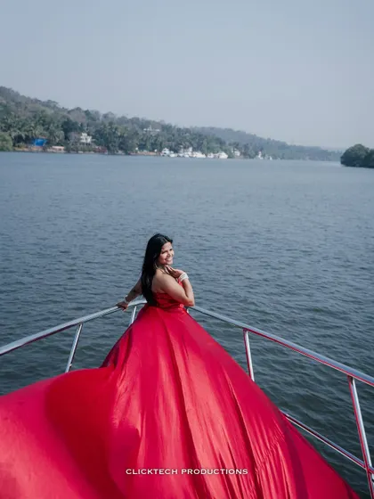 The bride-to-be looks back with a smile from the bow of a yacht, her red gown creating a stunning visual against the water and shoreline.