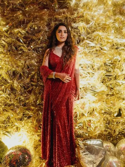 The bride stands radiant in her shimmering red Sangeet outfit against a textured gold foil backdrop, looking every bit the star of the evening.