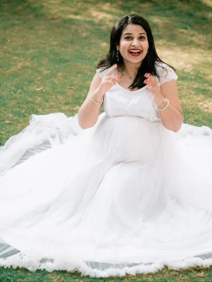 A fun and expressive moment from a park photoshoot. The mother-to-be, seated on the grass in a beautiful white gown, shares a joyful, candid laugh.