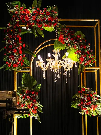 A beautiful decor element combining a golden arch, a mini chandelier, and lush arrangements of red flowers and tropical leaves, adding elegance to the stage design.