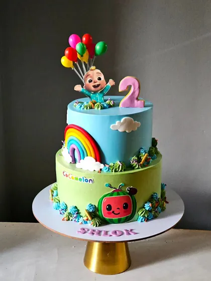 A cheerful two-tiered Cocomelon cake. This design features JJ with balloons on top and the Cocomelon logo on the side, surrounded by colorful details.