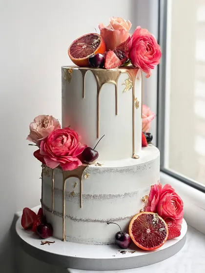 A stunning two-tiered cake combining a semi-naked finish, a gold drip, and an arrangement of fresh flowers and fruits like blood oranges and cherries.