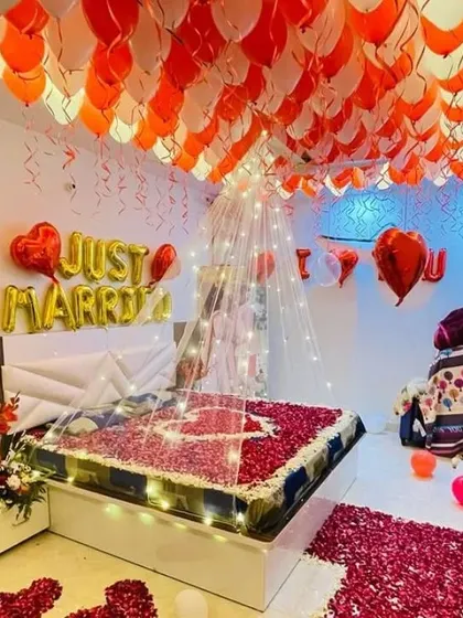 I specialize in creating memorable 'Just Married' room decorations. This setup includes hundreds of red and white balloons covering the ceiling, foil letter balloons, a light-up canopy over the bed, and a floor covered in artificial rose petals.