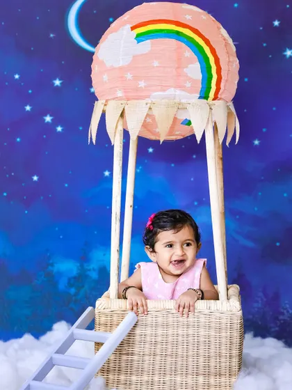 A happy baby girl peeks out from a hot air balloon basket prop. The starry night background and a rainbow on the balloon add to the magical, dreamy feel of the photoshoot.