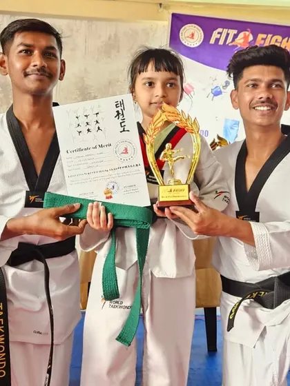 A young girl proudly holds her trophy, certificate, and new green belt, flanked by her senior coaches. Each belt earned is a victory.