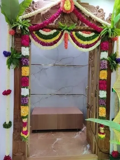 A pooja room door decorated with colorful flower garlands and banana leaves at the top, a classic combination for any auspicious occasion.