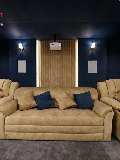 A full view of a 7.1.2 luxury home cinema in Karur, featuring a large sofa and two recliners in a warm, inviting fabric. The wood-paneled accent wall adds a touch of natural texture.
