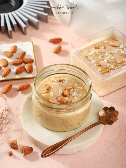 A jar of creamy almond dessert, styled with whole almonds and a decorative spoon on a pink surface.
