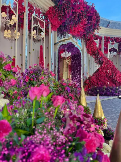 A view of the entrance, where a cascade of red flowers flows down the side of a mirrored arch, surrounded by a garden of purple blooms.