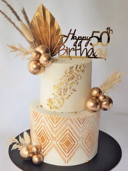 A stunning white and gold two-tier cake for a 50th birthday. The bottom tier has a geometric gold stencil, and the cake is adorned with dried florals and gold spheres for a modern, celebratory feel.