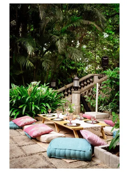 A beautiful low-table picnic arrangement on a stone patio, surrounded by lush greenery. The mix of pink and blue cushions adds a soft pop of color.