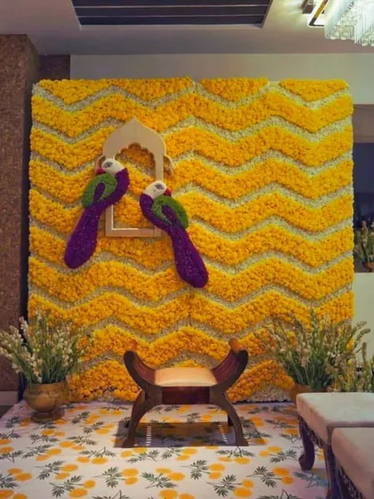 A creative Haldi photo booth with a chevron-patterned marigold wall and decorative parrots.
