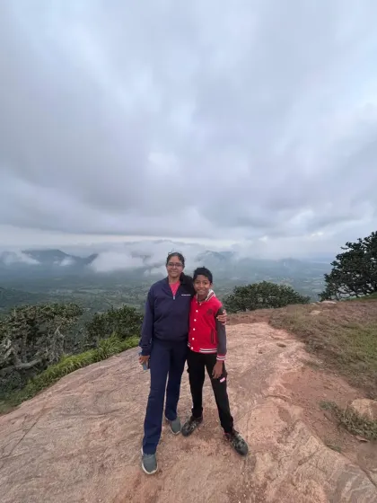 A mother and son duo smiling for a photo at the Kabbaldurga summit on a cloudy day. We welcome adventurers of all ages to create special memories together.
