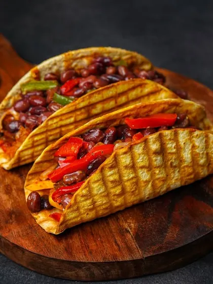 Two grilled tacos filled with beans and peppers, presented on a rustic wooden board with a side of dip.
