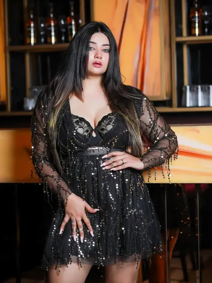 A model in a black sequined party dress with sheer sleeves, posing in a dimly lit bar setting.