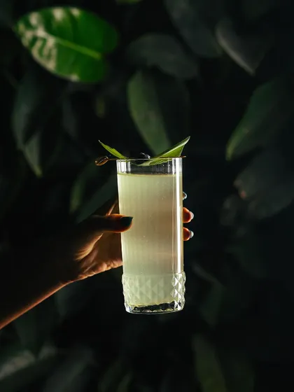 A hand holds a tall, elegant pandan-infused cocktail against a lush green background.