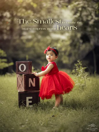 A beautiful first birthday portrait of a little girl in a vibrant red tulle dress, standing in a sunlit park next to rustic "ONE" blocks. This outdoor milestone session captures the magic of childhood.