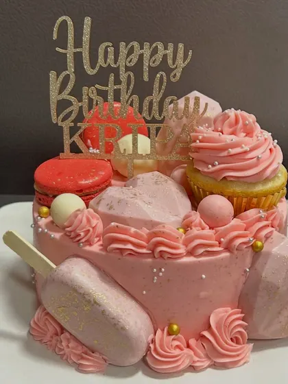 A fun and funky pink cake loaded with treats! It's topped with a cupcake, macarons, and geometric chocolate hearts for a truly indulgent birthday celebration.