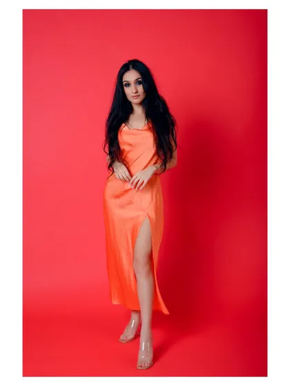 A vibrant, full-length fashion shot against a solid red background. The color contrast makes the orange dress pop, creating a bold and eye-catching image for a lookbook or campaign.