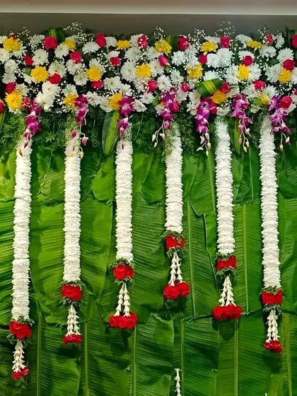 A close-up of a traditional backdrop made with fresh banana leaves. The top is adorned with a colorful mix of flowers and long jasmine garlands, creating a fresh and auspicious look.