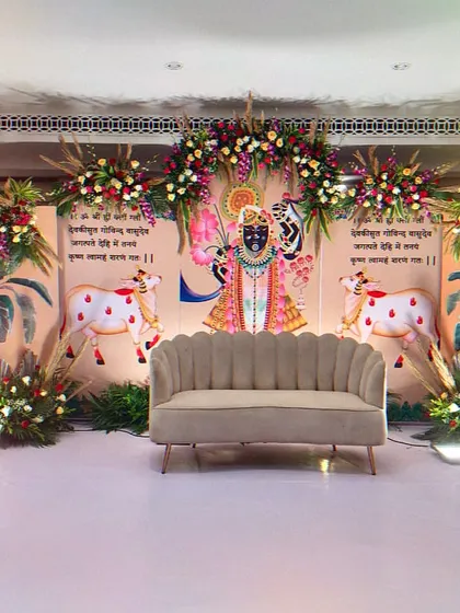 A beautiful stage designed for a religious ceremony, featuring traditional Pichwai art of Shrinathji and his cows. The decor is complemented by floral arrangements and banana leaf motifs, creating a serene and spiritual ambiance.