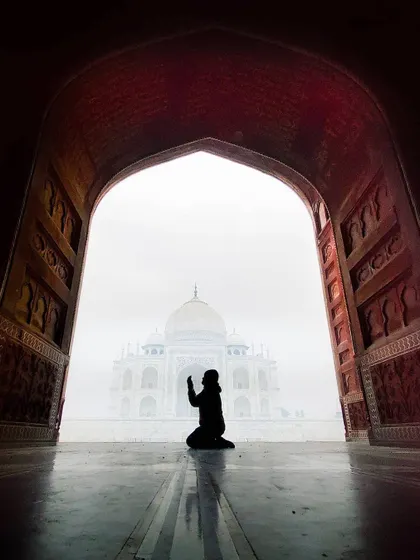 A person kneels in prayer, silhouetted within a grand archway that perfectly frames the distant Taj Mahal. This powerful composition uses framing to create a sense of reverence and scale.