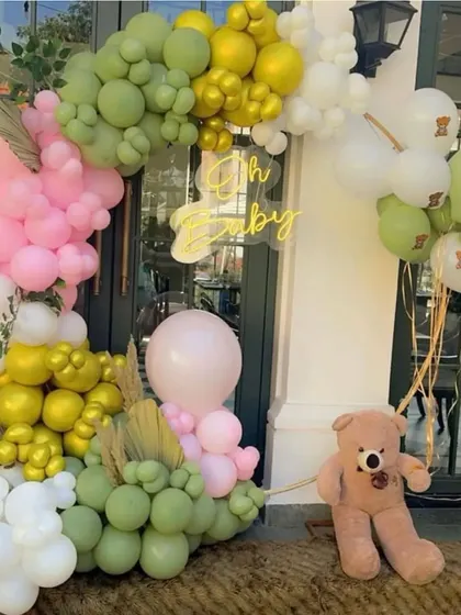 A close-up of a stylish "Oh Baby" shower decoration. This setup features a trendy color palette of olive green, pink, and white balloons, a neon sign, and a cute teddy bear prop.