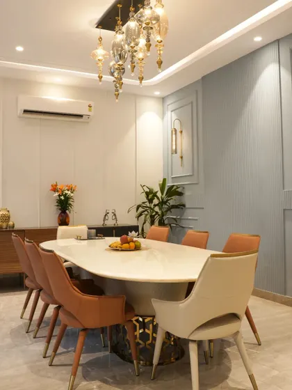 This dining area in a Noida project uses a pastel blue and grey color scheme with classic wall paneling. Tan and beige chairs add warmth around the marble-top table.