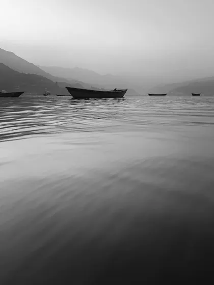 A few boats rest on calm, rippling water under a hazy sky. This minimalist and serene black and white photograph focuses on the subtle textures of the water and the quiet atmosphere.