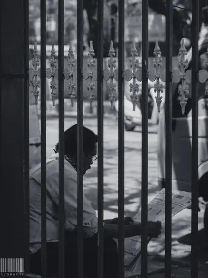 A man reading a newspaper, seen through the bars of a gate. This composition adds a layer of separation, making the viewer feel like a secret observer of a quiet moment.