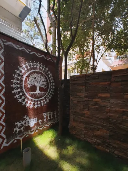 My Tree of Life mural on a compound wall, seen in the daylight. The sunlight reveals the texture of the wall and the clean, white pigment of the Warli painting, which includes a bullock cart scene below the central motif.