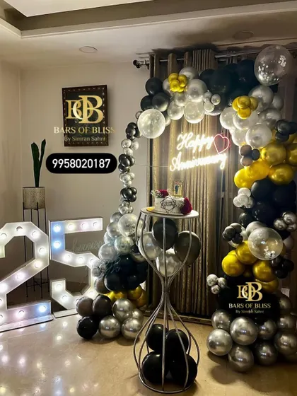 A classic and elegant 25th-anniversary decor for an intimate home celebration. The setup includes a balloon arch in black, silver, and gold, a neon sign, and marquee numbers.