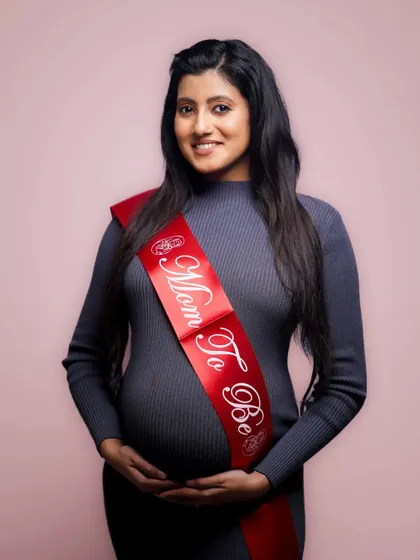 A smiling mother-to-be poses for a studio maternity photo against a soft pink background. This is a clean and modern style of maternity photography that I offer.