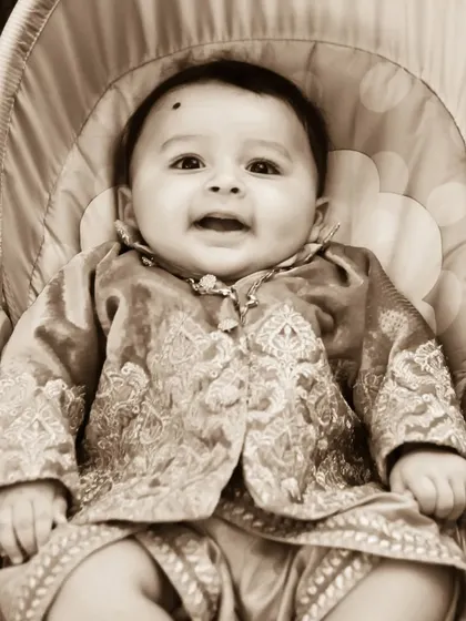 A sepia-toned portrait of a happy baby in a traditional embroidered outfit, sitting comfortably in a baby chair. This classic editing style adds a timeless feel to baby photos.