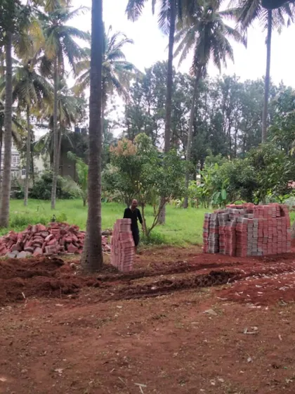 A wide shot of the farm garden site, with piles of red and grey pavers ready to be laid. The scale of the project is evident, with numerous coconut and mango trees in the background.