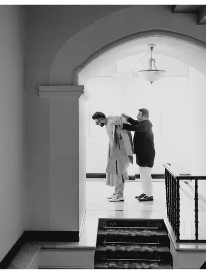 A candid, black and white photo of the groom getting ready with the help of a family member.