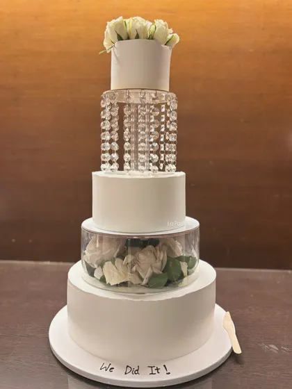 A five-tier masterpiece for a grand wedding celebration. This cake combines classic white tiers with modern clear separators filled with white roses and hanging crystals for a truly luxurious and breathtaking effect.