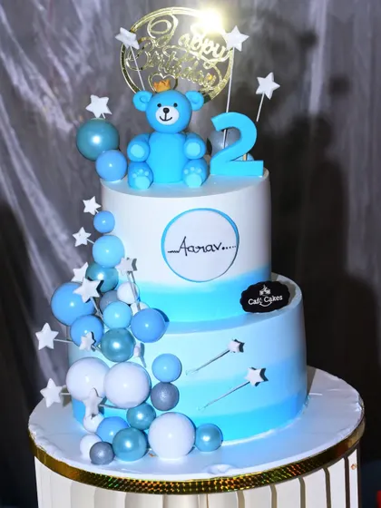 A two-tiered cake for a second birthday, decorated with a teddy bear and stars. The blue and white colors match the pastel theme of the party.