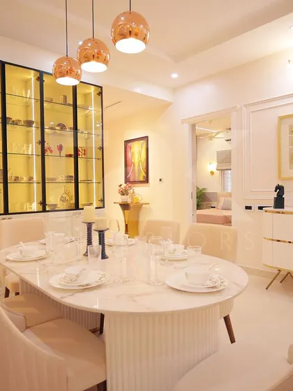 This elegant dining room features a white marble-top table and plush upholstered chairs. A statement crockery unit with gold-tinted glass and internal lighting adds a touch of glamour to the space.