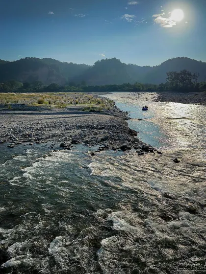 The sun shines brightly over the flowing river and rocky banks in the Himalayan foothills. You can almost feel the warmth of the sun and hear the sound of the water.