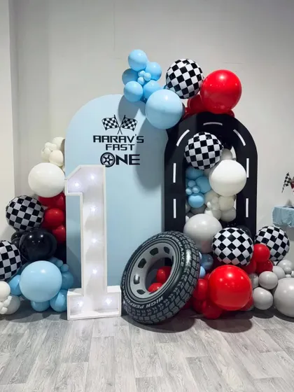 A "Fast One" first birthday setup with a race car theme. A large light-up number "1" is the centerpiece, surrounded by balloons and racing-themed props.