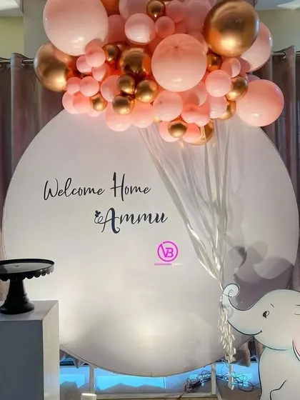 A simple and elegant welcome home backdrop for Ammu. The design features a clean white circular backdrop, a pink and gold balloon arrangement, and a cute elephant cutout.