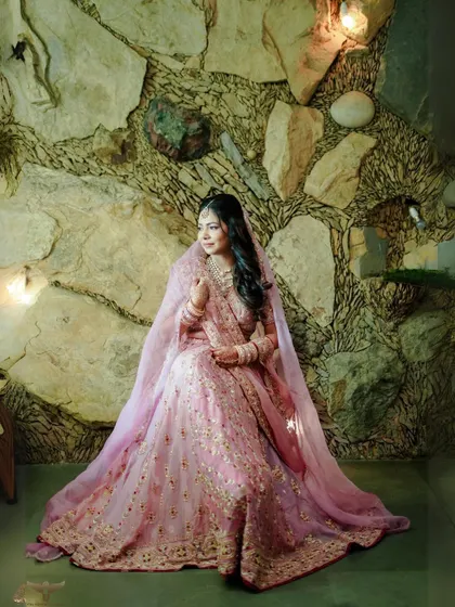 The bride is seated like royalty in this full-length portrait. Her voluminous pink lehenga spreads out beautifully against the rustic stone wall, creating a picture of grace.